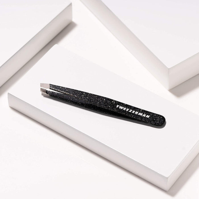 Anniversary Slant Tweezer | Tweezerman in black with a speckled design, displayed on a white geometric platform, ideal for precise eyebrow grooming in skincare routines.