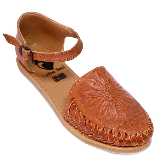 A tan leather flat sandal with an ankle strap, featuring a large embossed sunflower design on the toe box and braided detailing along the front edge,