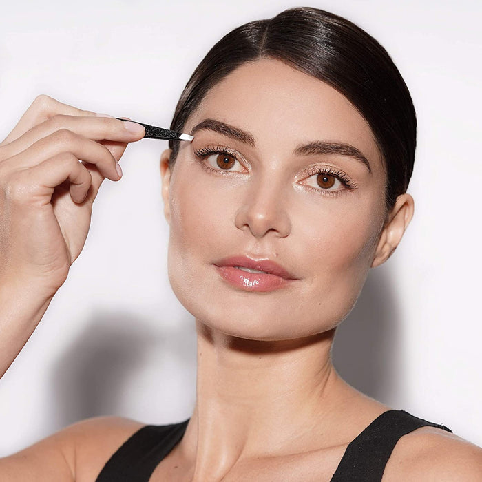 Woman with flawless skin using the Anniversary Slant Tweezer | Tweezerman to groom her eyebrows in a well-lit setting, highlighting a skincare and body care routine.