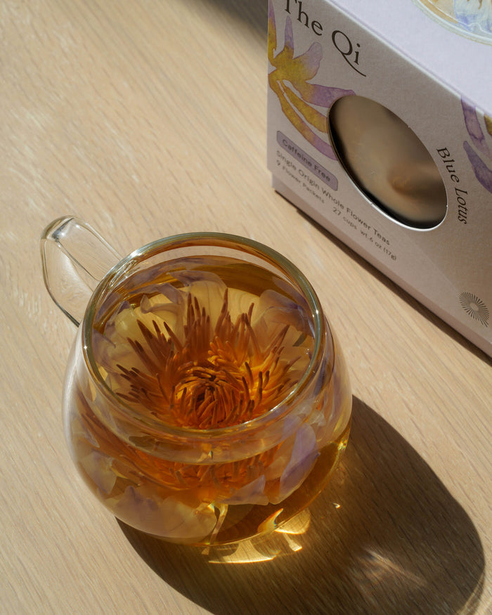 Glass cup of Blue Lotus Whole Flower Tea | The Qi with the flower steeped inside, placed next to its packaging on a wooden surface in natural light.