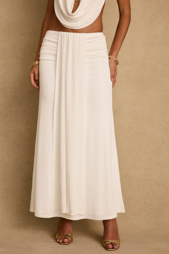 Emmeline white maxi skirt with ruched waist and front drape, styled with gold heels.