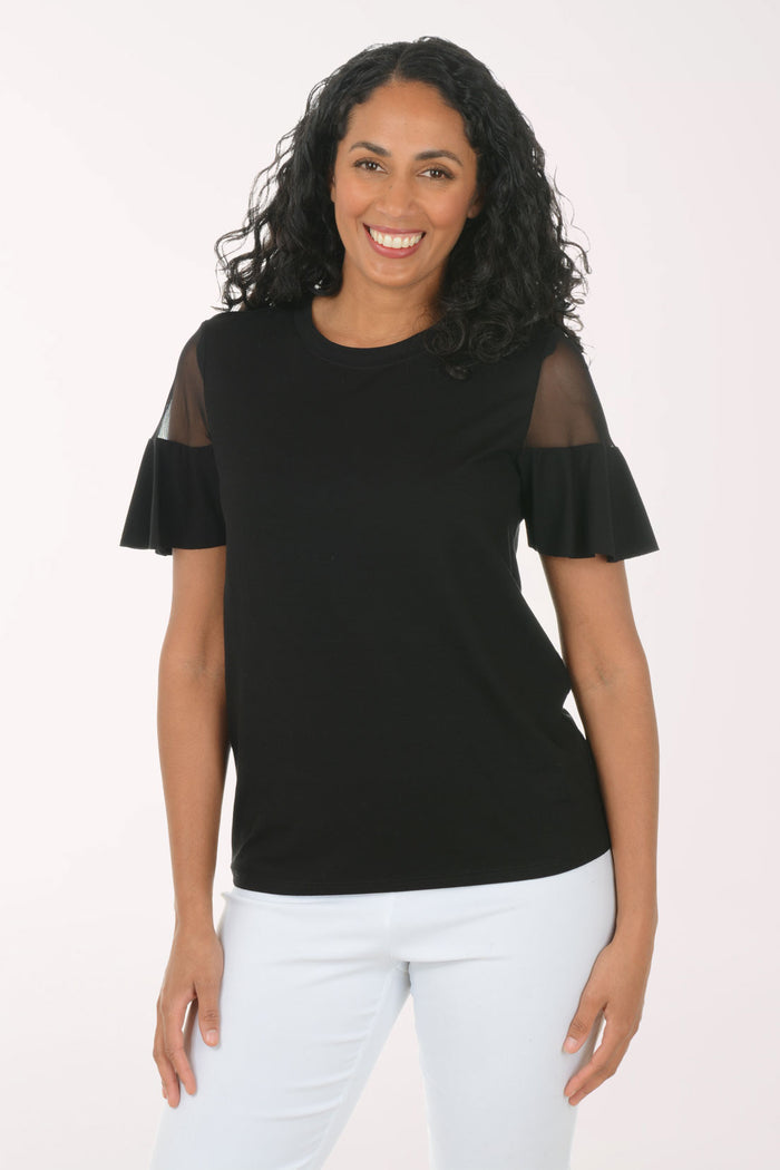 Woman wearing a black top with sheer sleeves and white pants on a light pink background