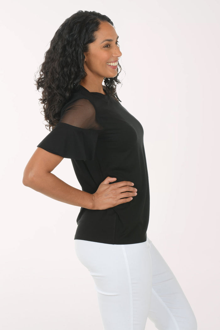 Woman wearing a black top with sheer sleeves and white pants on a light pink background