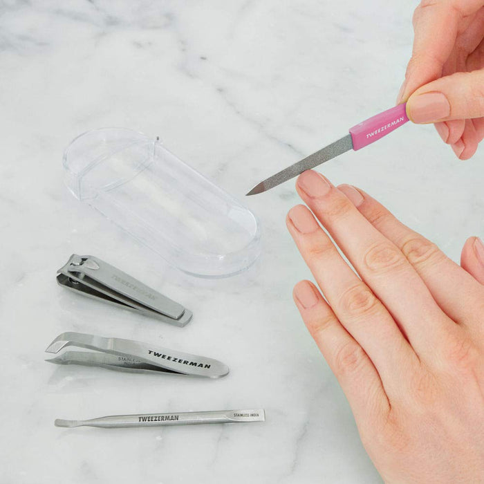 A person filing fingernails with a pink-handled file from the Mini Nail Rescue Kit | Tweezerman, shown with nail clippers, tweezers, and a cuticle pusher on a marble surface.