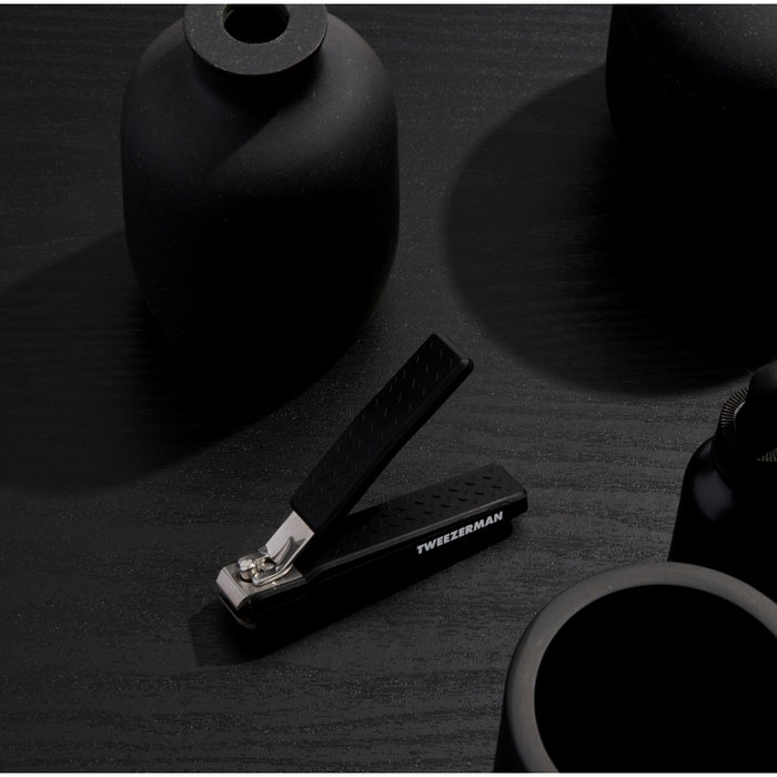 Precision Grip Toenail Clipper | Tweezerman with textured black handle for secure hold, placed on a matte black surface alongside minimalistic black decor items.