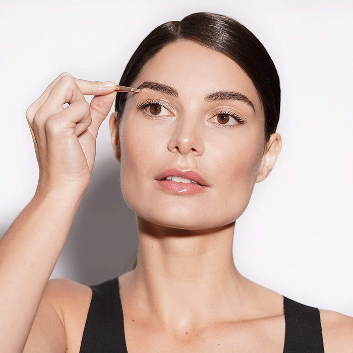 Woman holding the Rose Gold Mini Slant & Case | Tweezerman near her eyebrow, demonstrating precise grooming with a sleek makeup look and pulled-back hair.