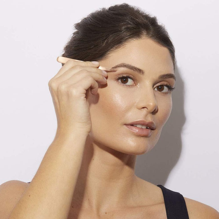 Woman using Rose Gold Slant Tweezer | Tweezerman to shape her eyebrows in a close-up beauty portrait with natural makeup and glowing skin.