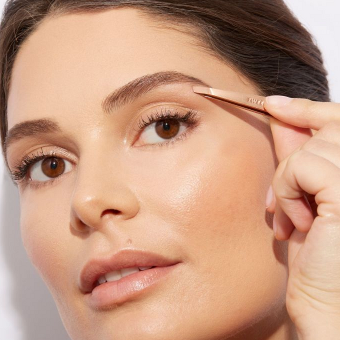 Close-up of a woman using the Rose Gold Slant Tweezer | Tweezerman to groom her eyebrow, highlighting precision and skincare beauty tools.