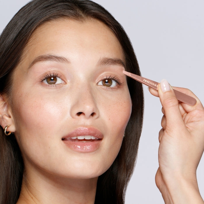 Close-up of a woman using the Rose Gold Slant Tweezer | Tweezerman to groom her eyebrows, highlighting precision and beauty care in a skincare routine.
