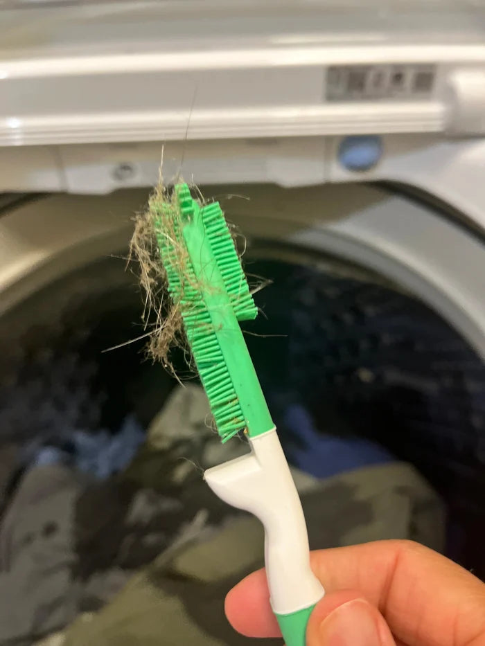 Green and white  silicone brush with hair residue held in front of a washing machine.